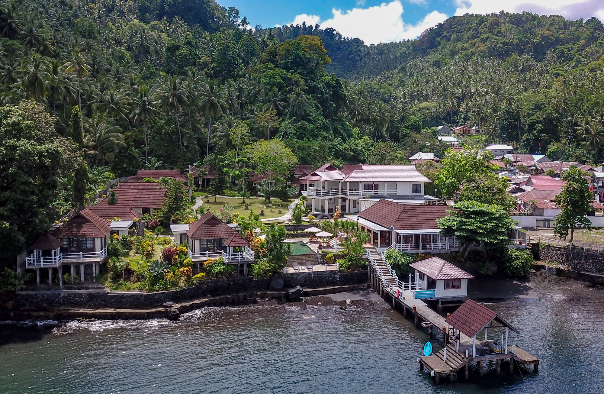 Aerial view of Solitude Lembeh Resort, showcasing villas, jetty, pool, lush gardens, and scenic green hills under a blue sky