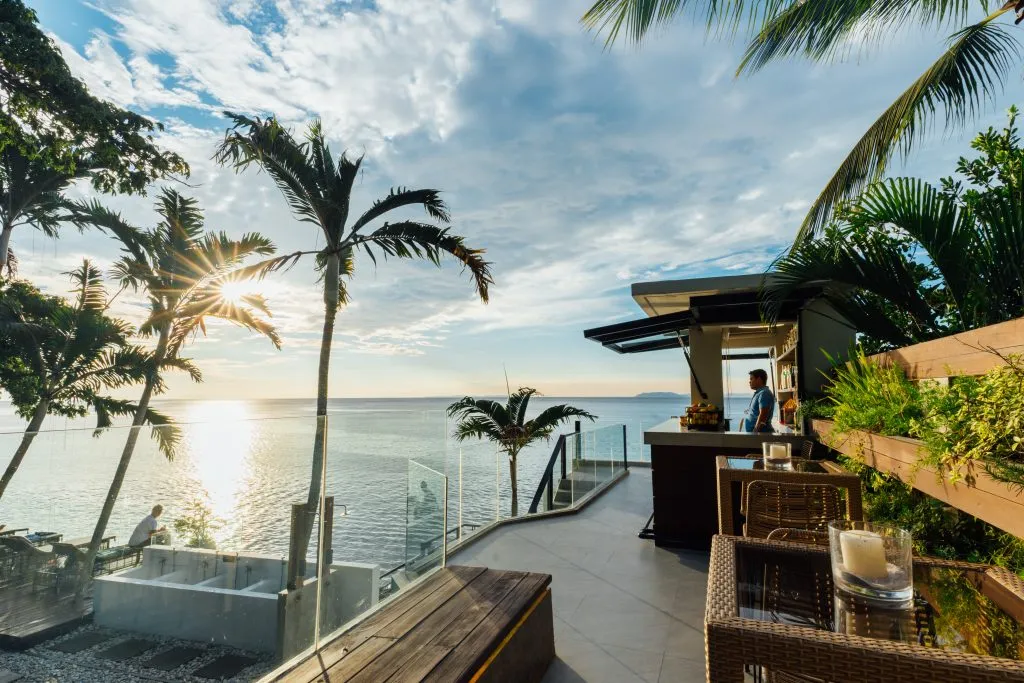 Oceanfront sunset bar terrace with palm trees and glass railing at Solitude Acacia Resort