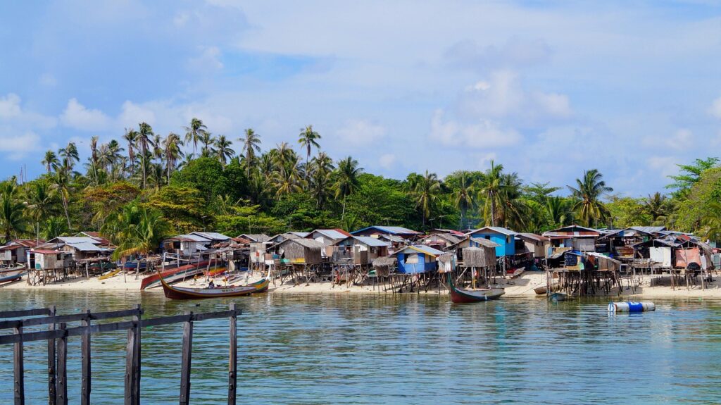 Traditional stilt houses and fishing boats along the shore of Mabul Island, surrounded by palm trees and lush tropical vegetation.