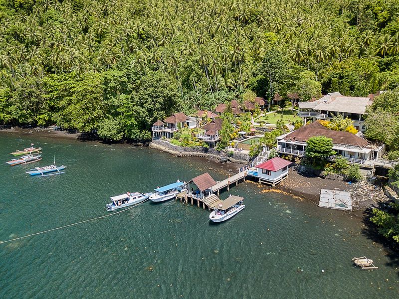 Aerial view of Solitude Lembeh Resort, a beachfront dive resort surrounded by lush tropical forest in Lembeh Strait, Indonesia, with dive boats docked at the private jetty.