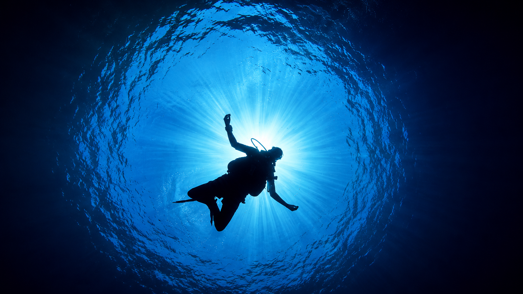 Silhouette of a scuba diver suspended beneath the surface with sunlight streaming through blue water, representing a diver returning to diving after a long break.