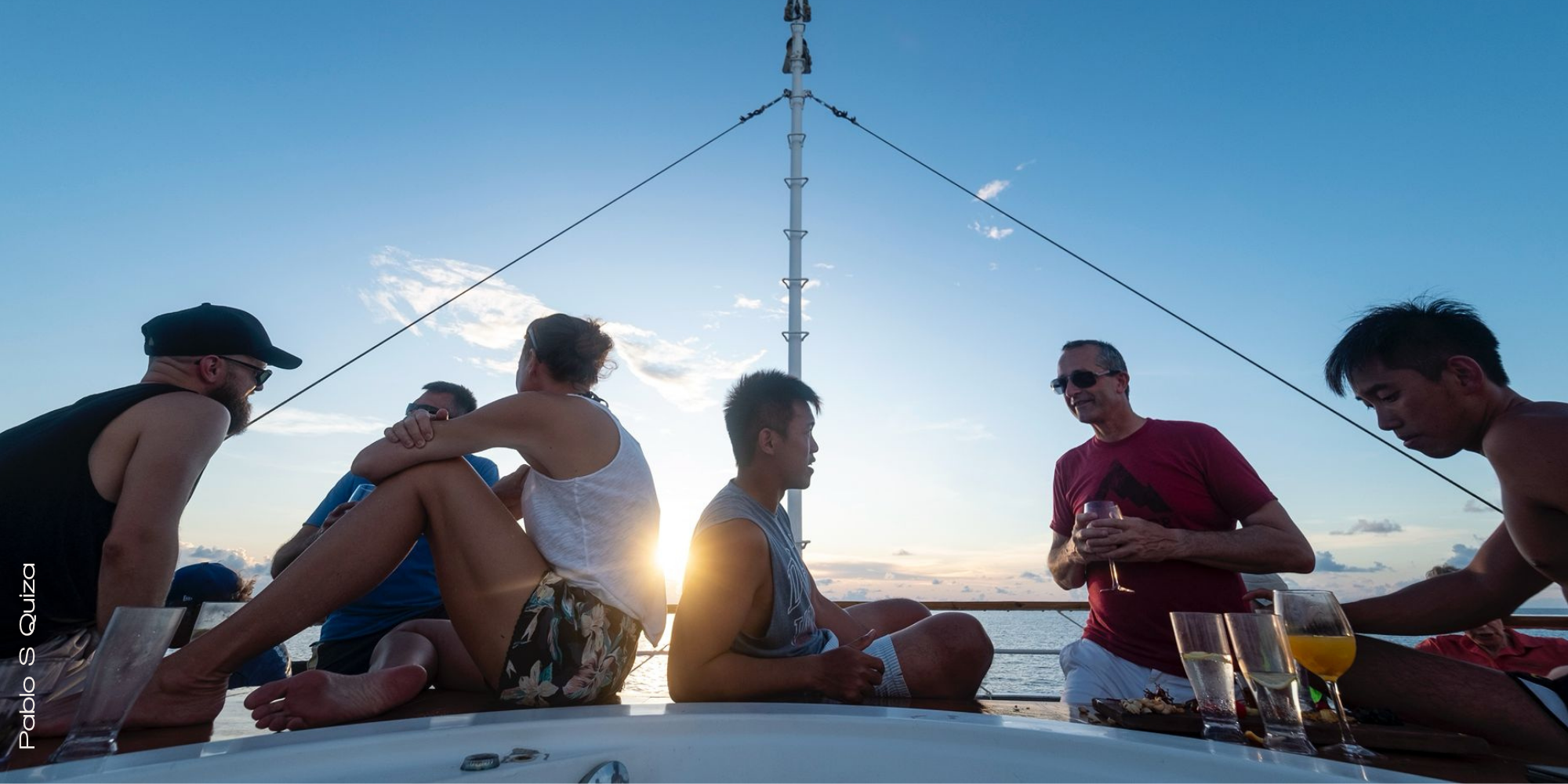 Group of scuba divers relaxing on a liveaboard sundeck at sunset, drinking coffee and laughing together after a day of diving, with the ocean and islands in the background.